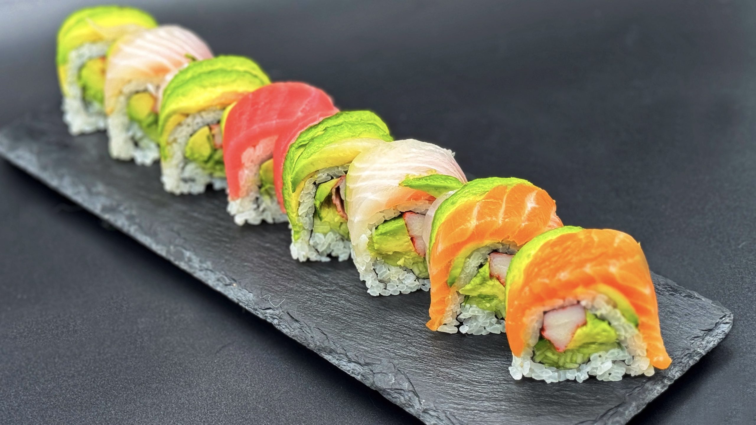 Rainbow sushi roll neatly sliced and arranged on dark slate platter, layered with fresh salmon, tuna, white fish and avocado over crab and vegetable filling inside rice.