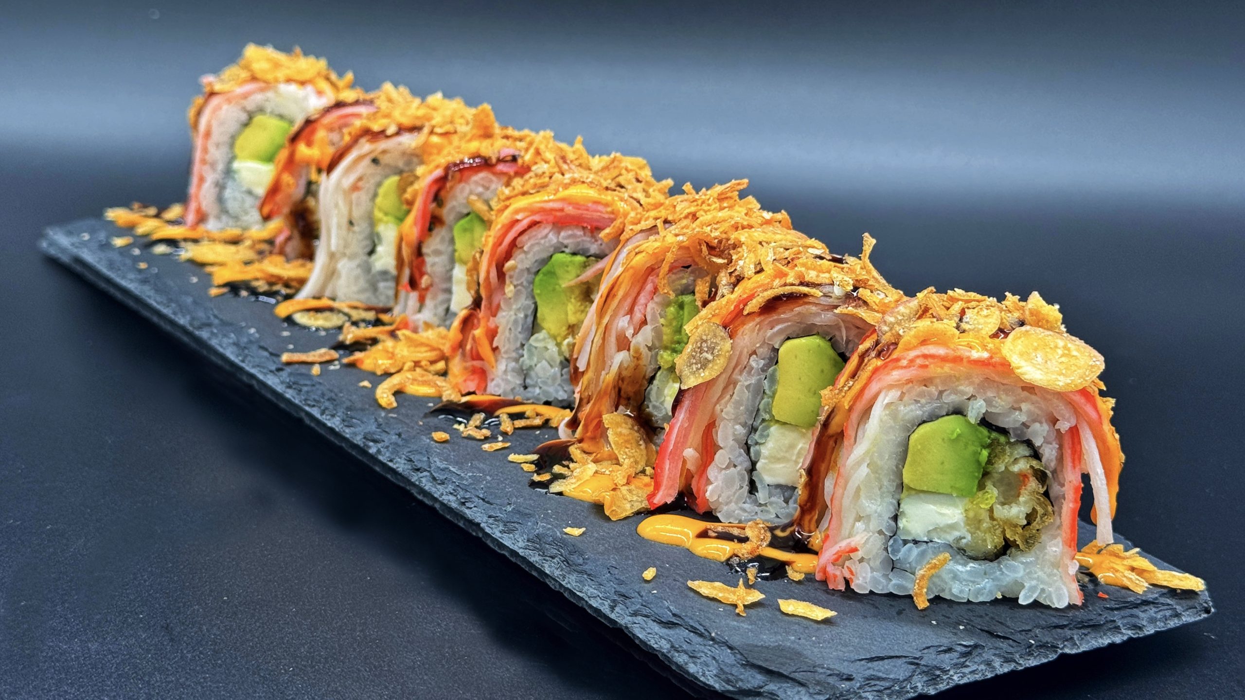 Sushi roll slices placed on dark slate plate, generously topped with crunchy fried onion flakes, drizzled with spicy mayo and dark eel sauce, avocado and cream cheese filling inside.