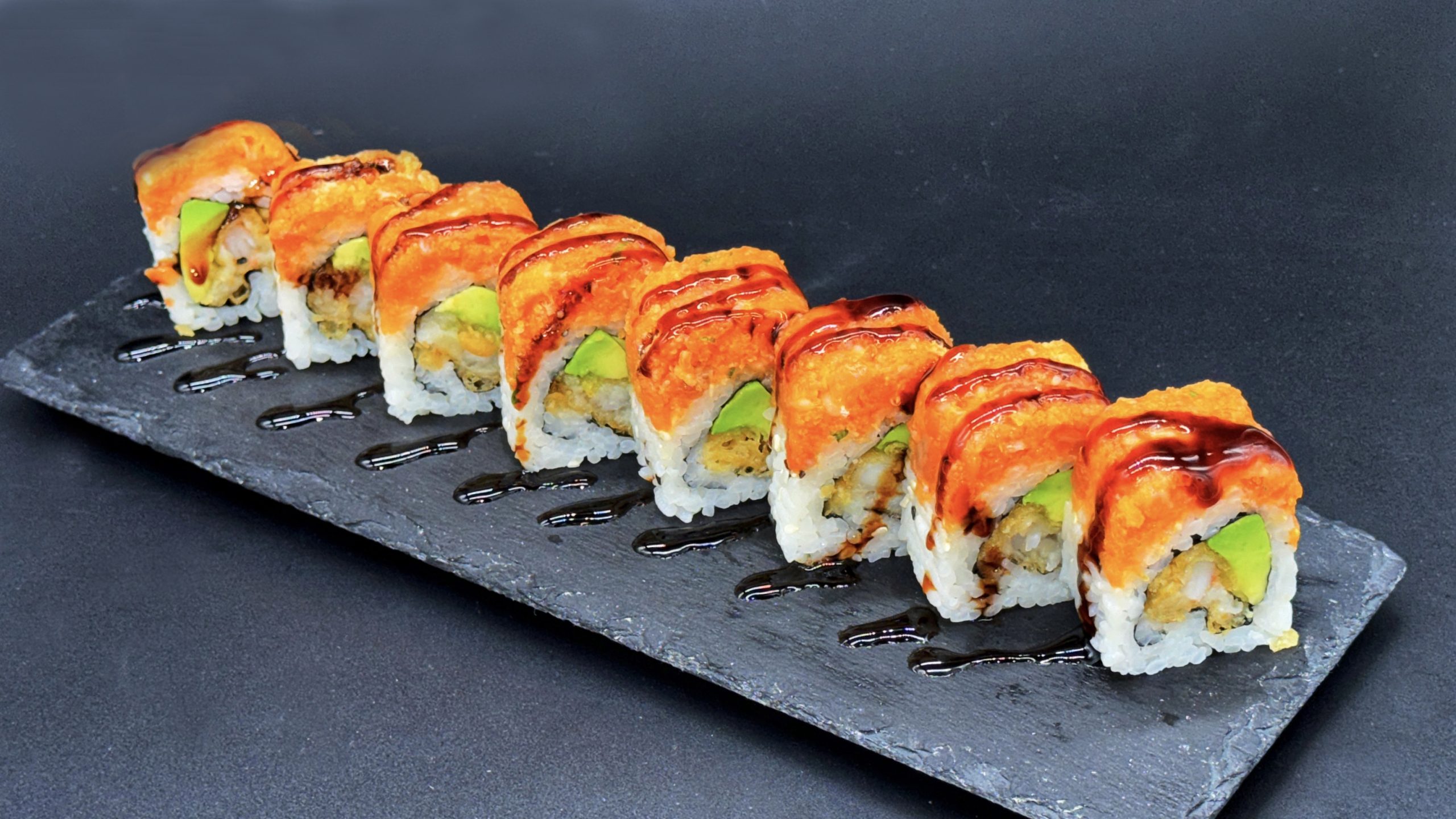 Sliced sushi roll pieces arranged on dark slate plate, topped with crispy orange tempura crunch, avocado inside, decorated with streaks of glossy dark eel sauce.