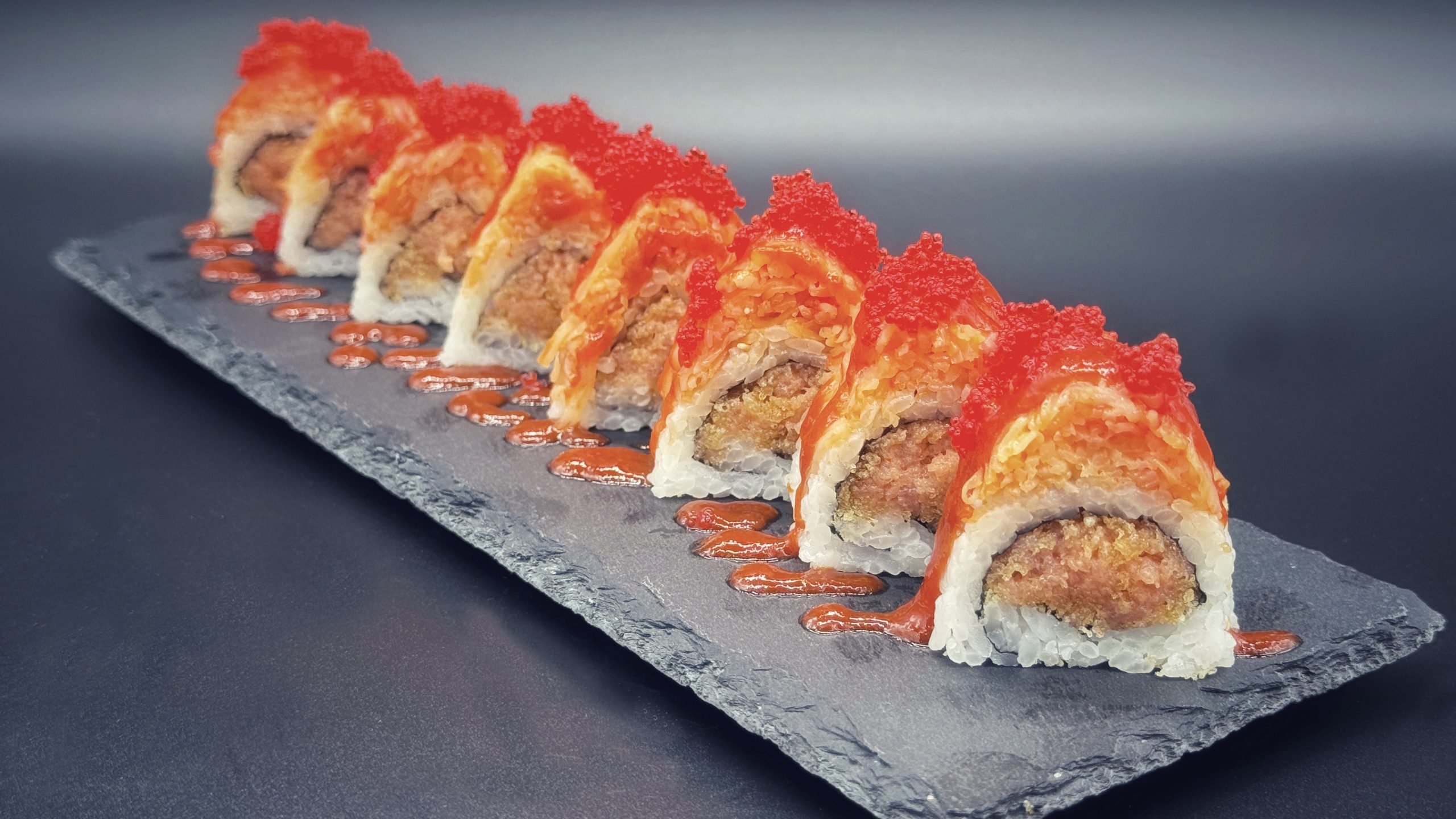 Sushi roll filled with spicy tuna, topped with bright red tobiko roe, vibrant red spicy sauce drizzled along dark slate plate, neatly arranged in straight row.