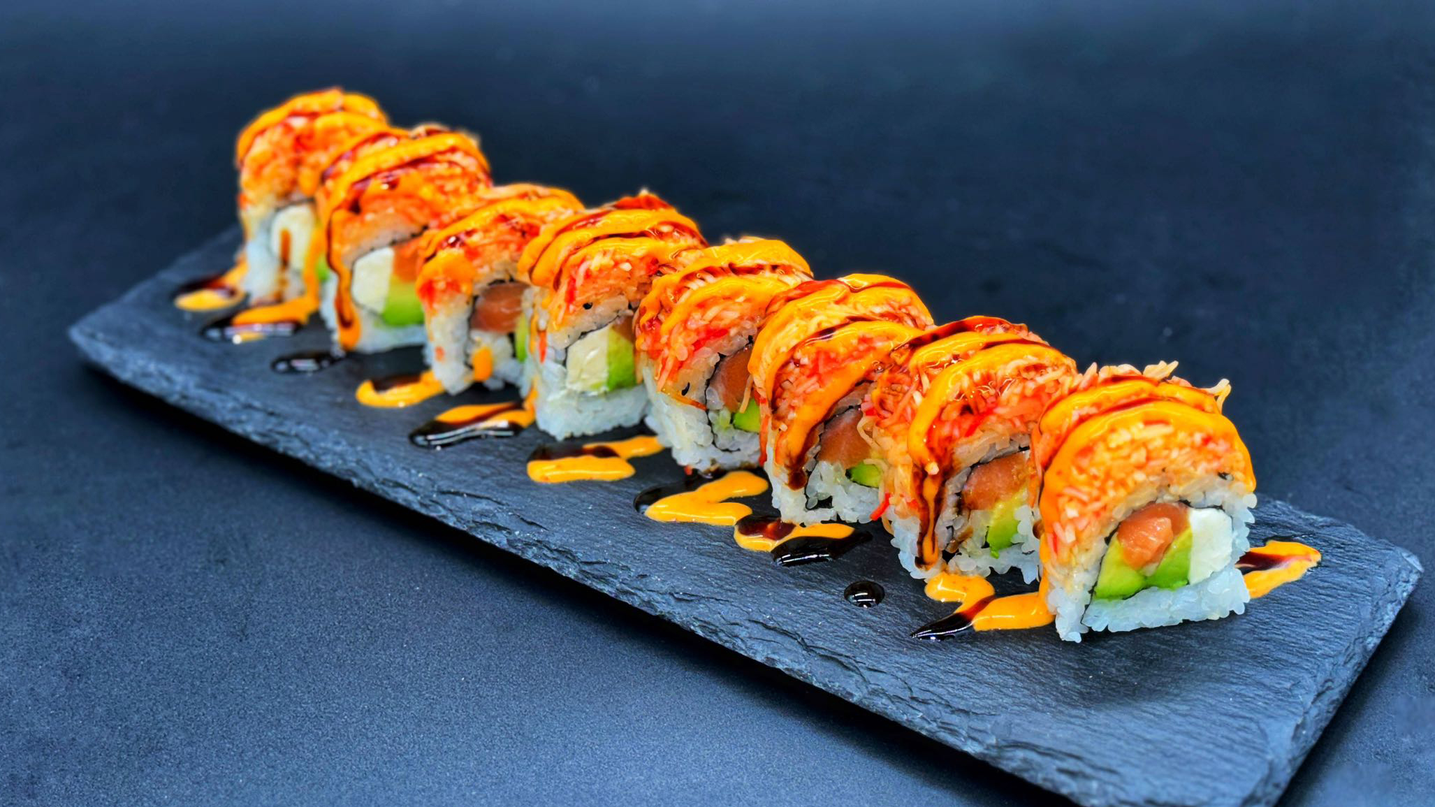 Eight pieces of specialty maki sushi roll arranged on dark slate platter, filled with salmon, avocado and cream cheese, topped with spicy crab meat, drizzled with orange spicy sauce and dark eel sauce against dark background