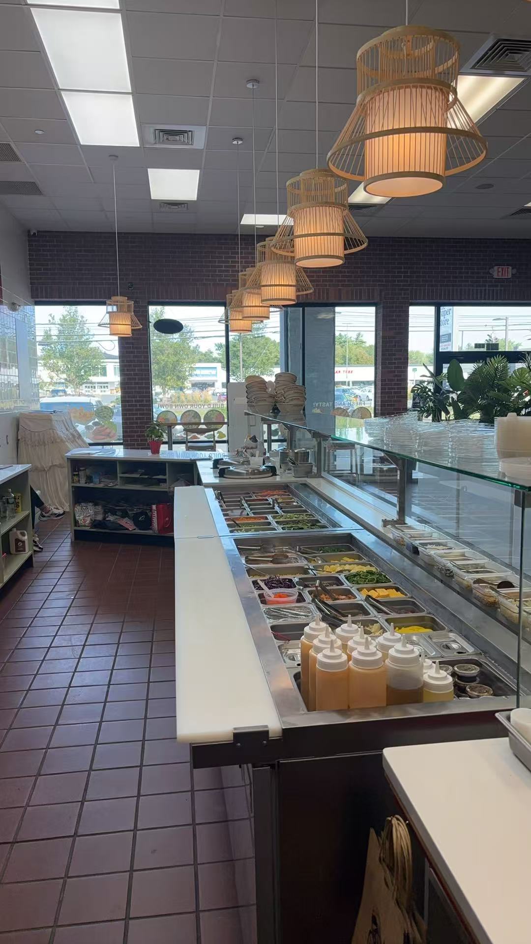 Poke-CO-2016-Bowl-Poke-Bowl-Restaurant-Bristol Self-serve poke bar with stainless steel trays of fresh ingredients, sauce bottles, and warm pendant lighting over a tiled floor.