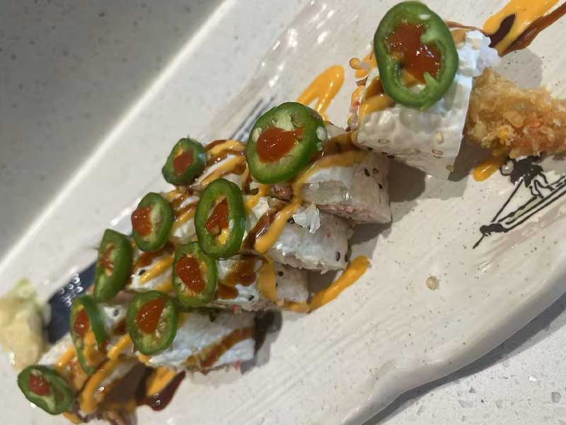 Sliced spicy sushi roll arranged on white ceramic plate, topped with fresh jalapeño slices, red chili sauce, drizzled creamy yellow and dark brown teriyaki sauces.