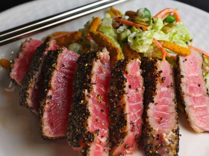 Sliced pepper‑crusted seared ahi tuna on white plate, pink rare tuna interior, side mixed fresh vegetable salad and metal chopsticks on plate edge.