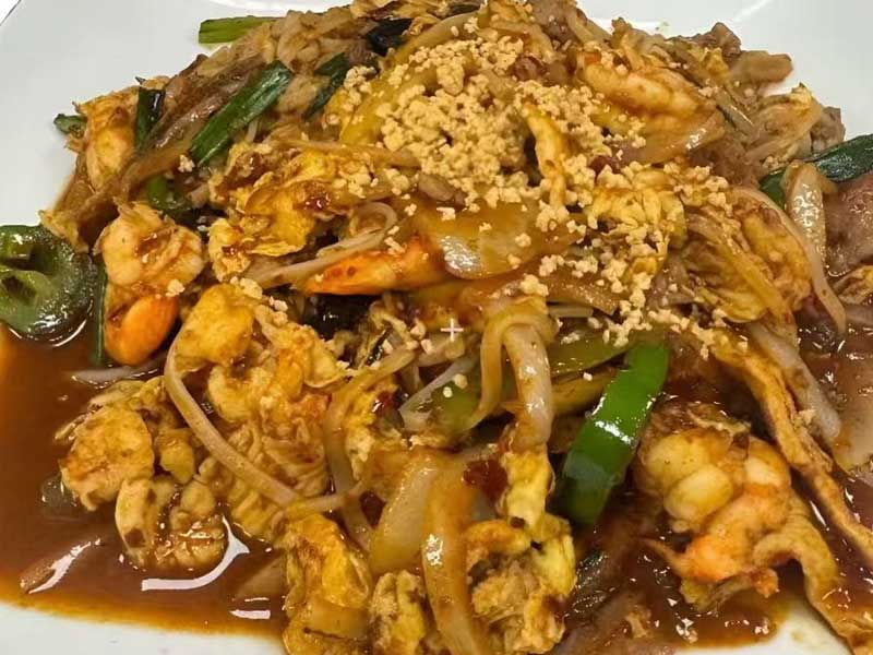 Stir‑fried rice noodles with shrimp, scrambled egg, green pepper, onion, crushed peanuts, coated in rich brown savory sauce, served on a white plate.