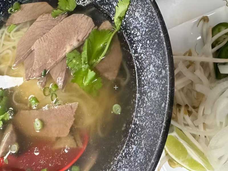 Beef pho noodle soup in speckled black bowl, sliced beef, cilantro and green onion in clear broth; side plate with bean sprouts, lime and chili.