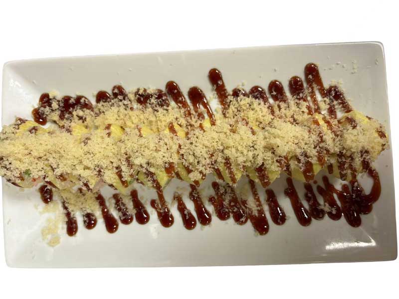 Top‑down shot of sliced sushi roll on rectangular white plate, generously sprinkled crispy crumbs, streaky dark brown teriyaki sauce drizzled across the whole dish.
