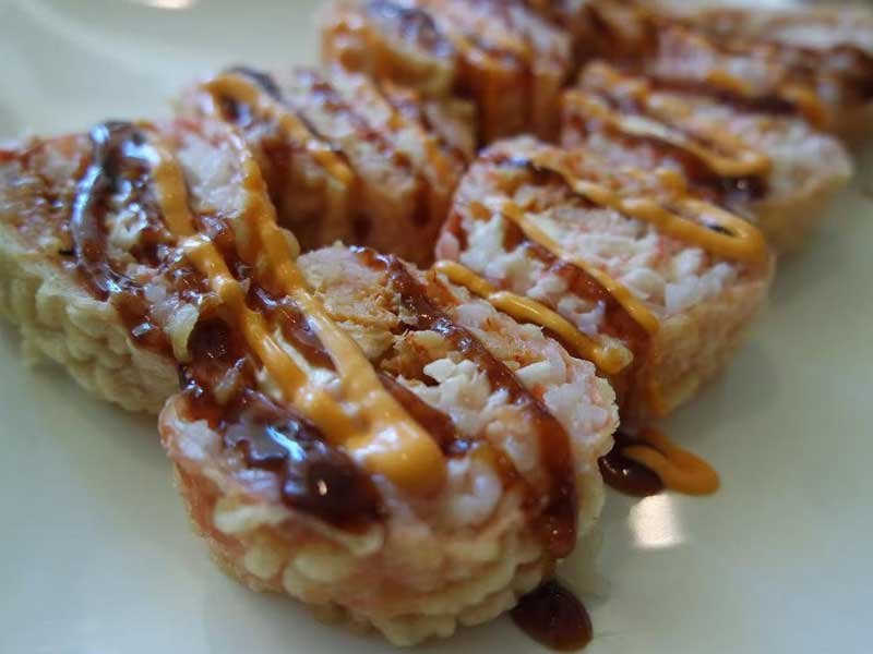 Close‑up view of sliced crab‑filled sushi rolls on white plate, dual drizzle of spicy orange cream sauce and thick dark brown teriyaki sauce over rice.