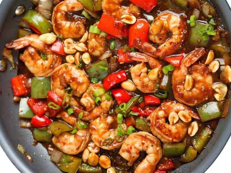 Top‑down view of kung pao shrimp in dark pan, plump cooked shrimp mixed with diced bell peppers, peanuts, green scallions in spicy glossy sauce.