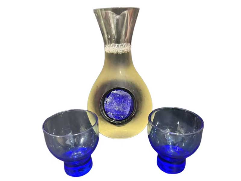 Glass sake carafe filled with pale yellow beverage with ice insert, paired with two matching deep‑blue glass sake cups against plain white background.