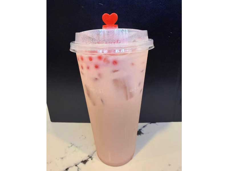 Tall transparent plastic cup with iced pink milky drink, red popping boba and ice cubes, red heart‑shaped stopper on plastic lid.