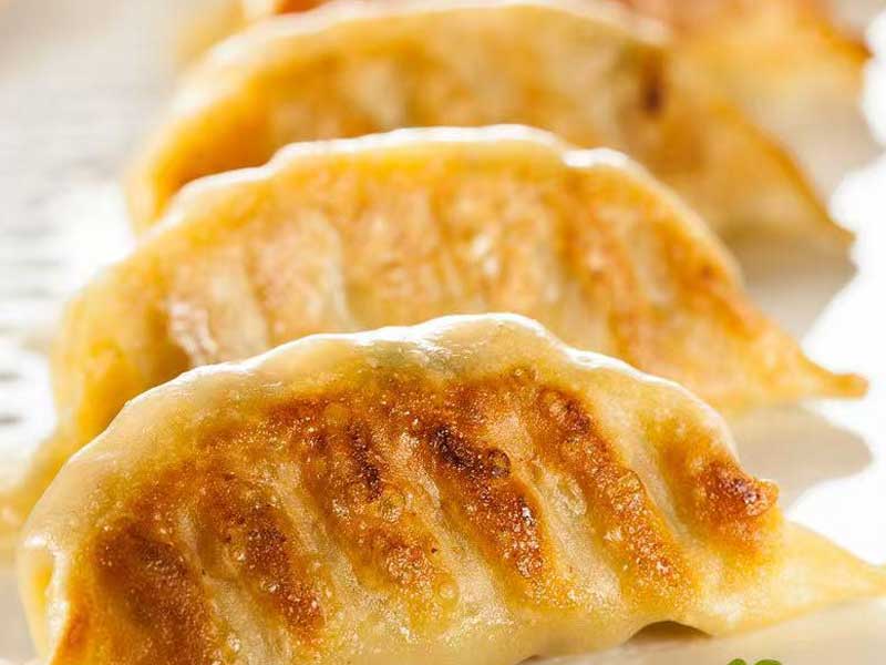Close‑up of pan‑fried gyoza dumplings on light plate, golden‑brown crispy seared bottoms, soft pale folded dumpling wrappers stacked in layers.