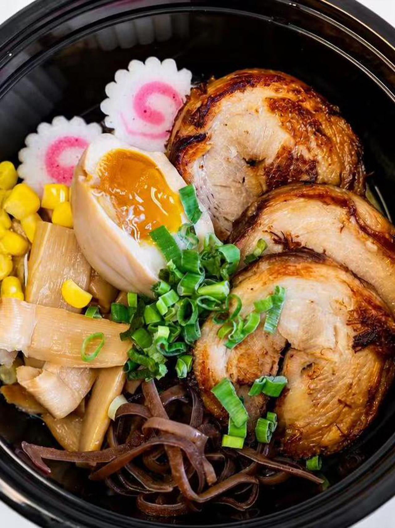 Ramen bowl with chashu pork, soft-boiled egg, corn, bamboo shoots, and scallions in a black container