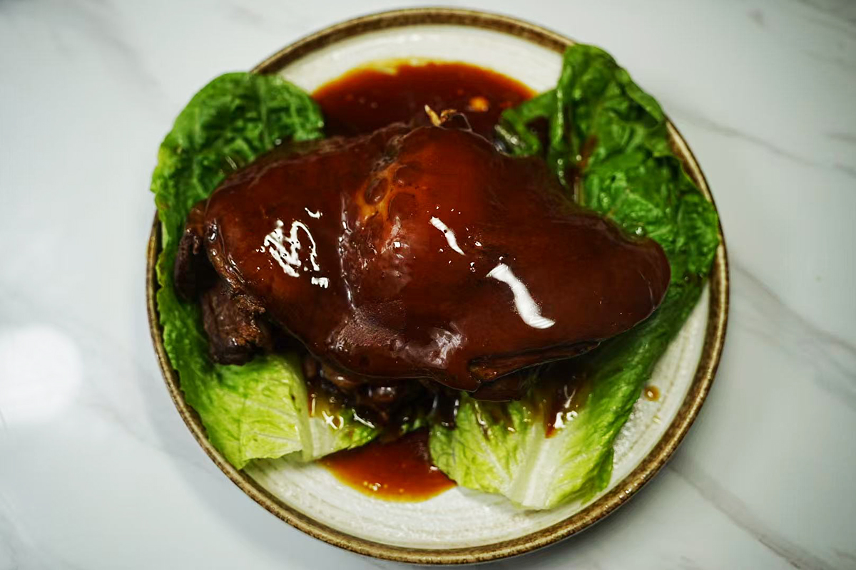 A whole braised pork hock with glistening dark amber skin, resting on a bed of fresh green lettuce, surrounded by rich brown sauce at Chef Ma，a Chinese Restaurant in Edison