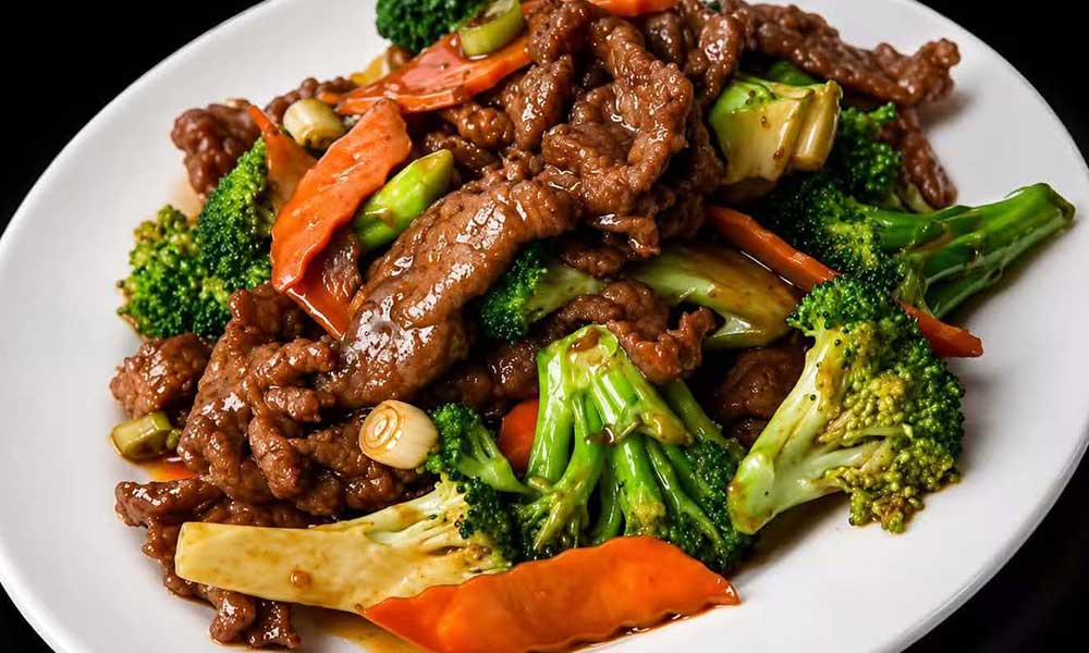 Glossy stir‑fried beef with fresh broccoli, carrot slices and green onion coated in rich brown sauce, served on a clean white plate against dark background.