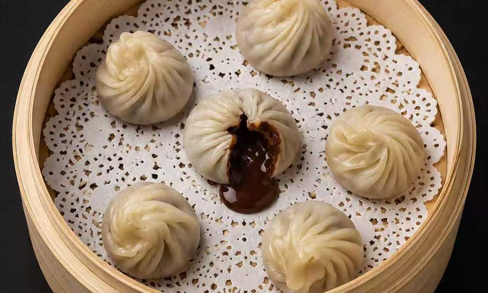 Six soup dumplings in bamboo steamer on lace paper, one dumpling split open with savory brown soup oozing out, top‑down food shot.