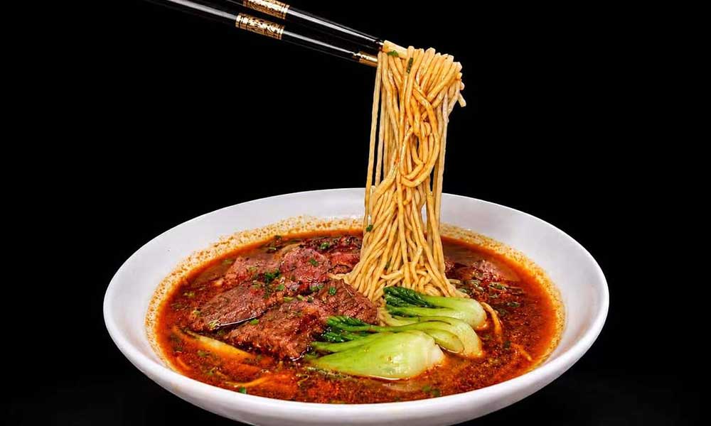 Spicy red beef noodle soup in white bowl, chopsticks lift up yellow noodles, tender beef slices and bok choy sit in rich reddish broth.