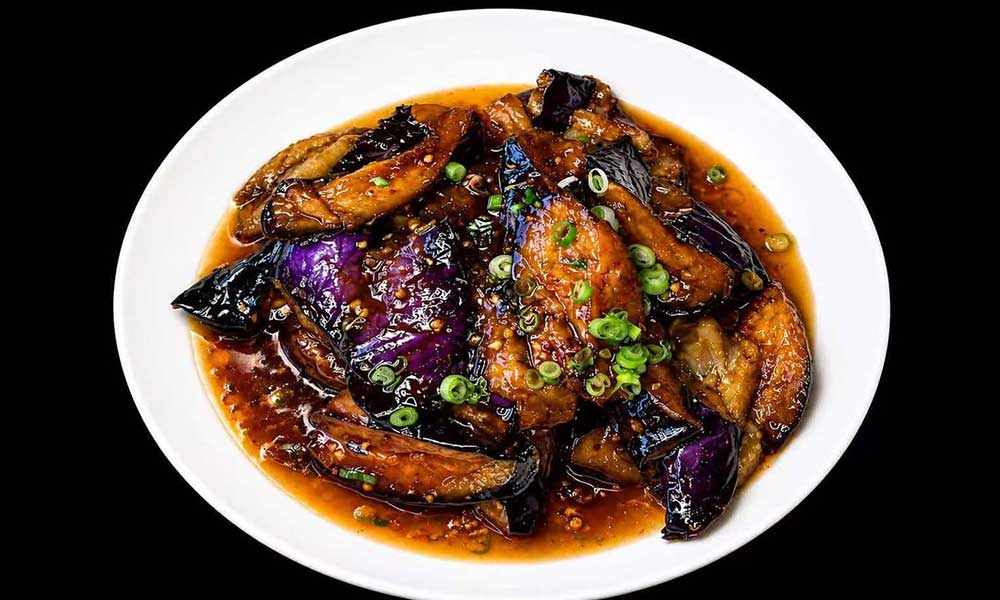 Braised eggplant chunks covered in glossy reddish‑brown savory sauce, garnished with chopped green scallions, presented on a plain white plate.