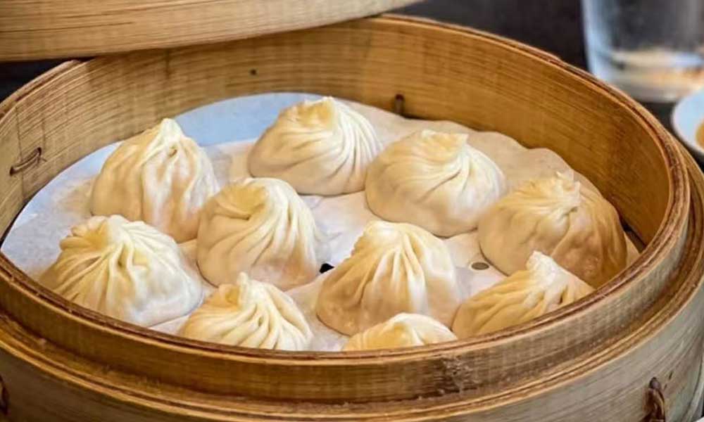 Multiple pleated soup dumplings neatly stacked inside open bamboo steamer lined with parchment paper, soft pale dough with twisted tops.