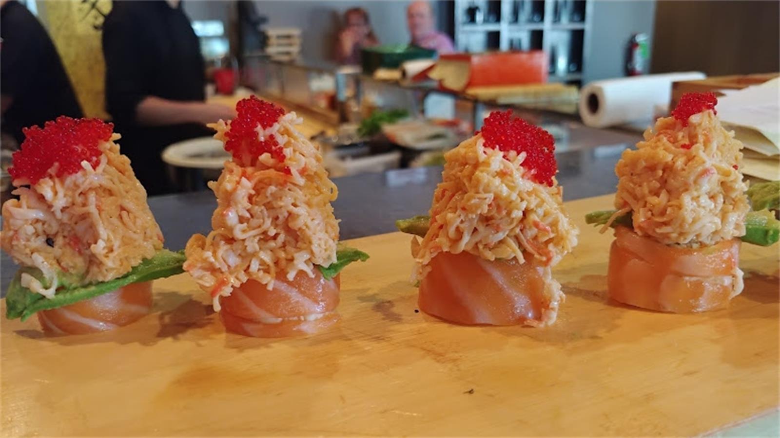 Four salmon wrapped sushi bites topped with crab salad and bright red tobiko roe on wooden board at Cho Sushi Japanese Fusion, a Japanese restaurant in Austin