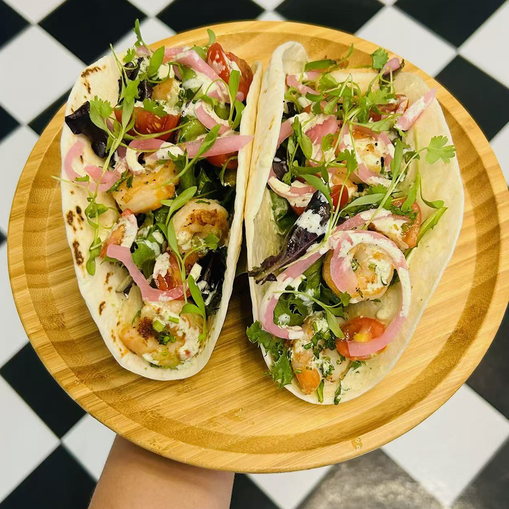 Two shrimp tacos with pickled onions, greens, and sauce on a wooden plate at Costa，a Colombian Restaurant in Houston