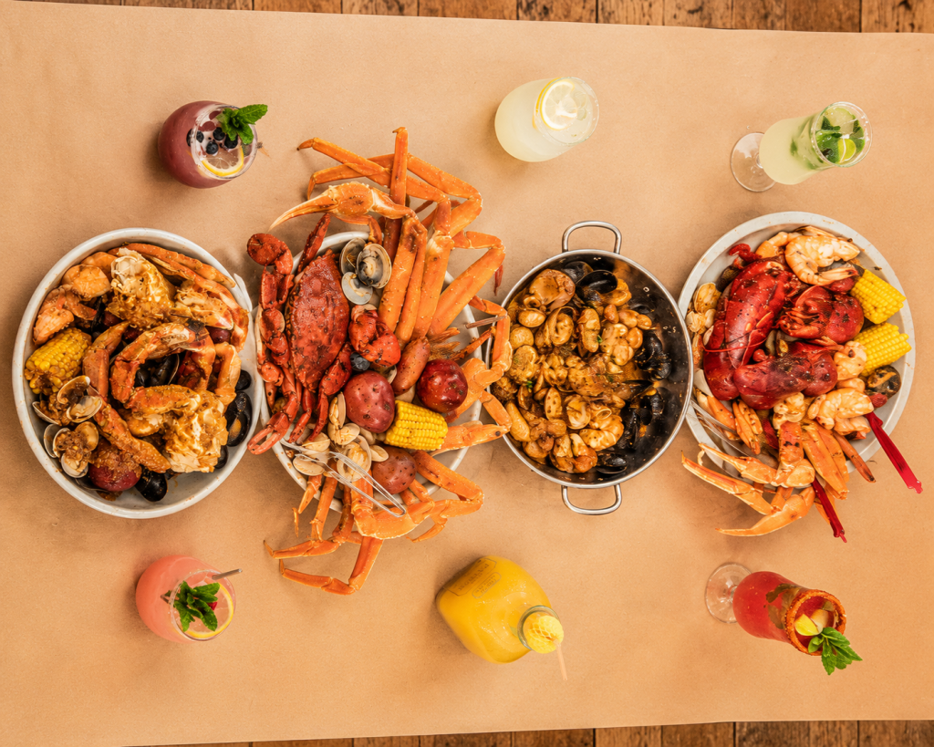 Overhead view of Cajun seafood boil platters with crab legs, lobster, mussels, shrimp, corn and drinks served on brown paper