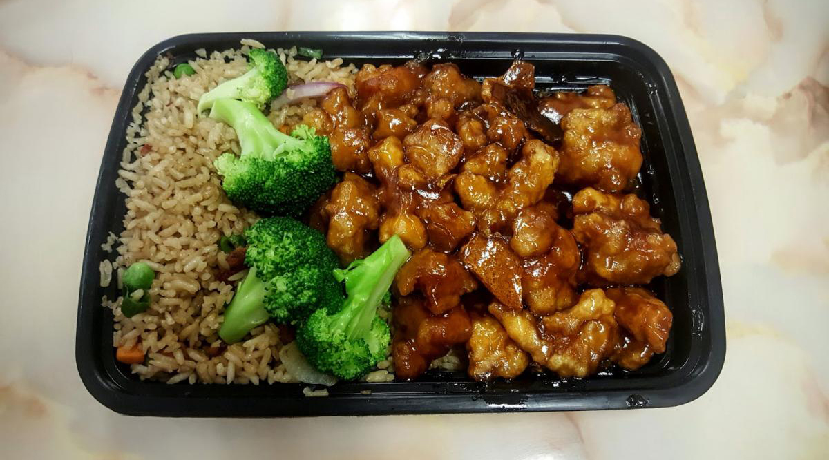 Black plastic takeout tray with General Tso's chicken in shiny sauce, fried rice and fresh broccoli florets.