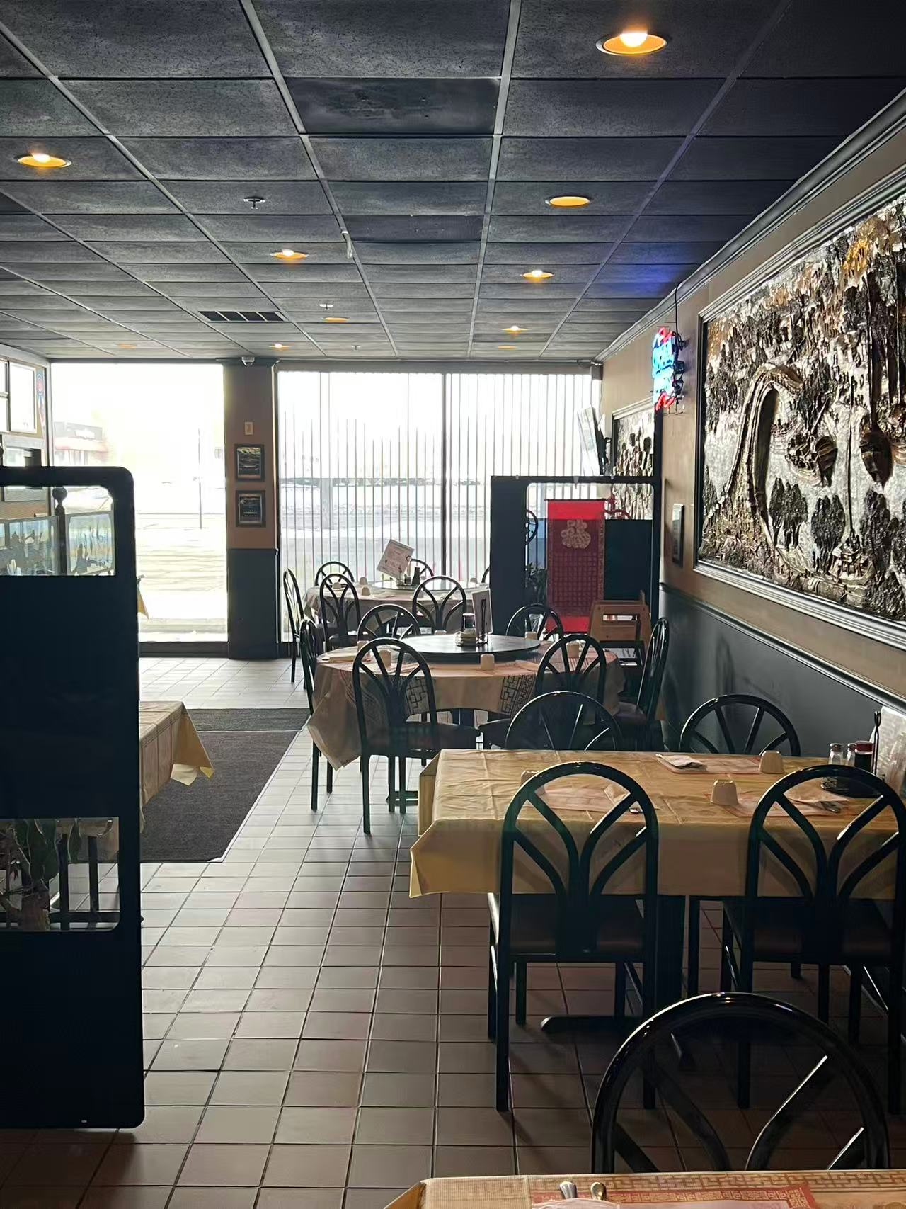 Empty restaurant interior: tiled floor, round tables with beige cloths, black chairs, large wall mural, and windows with vertical blinds.