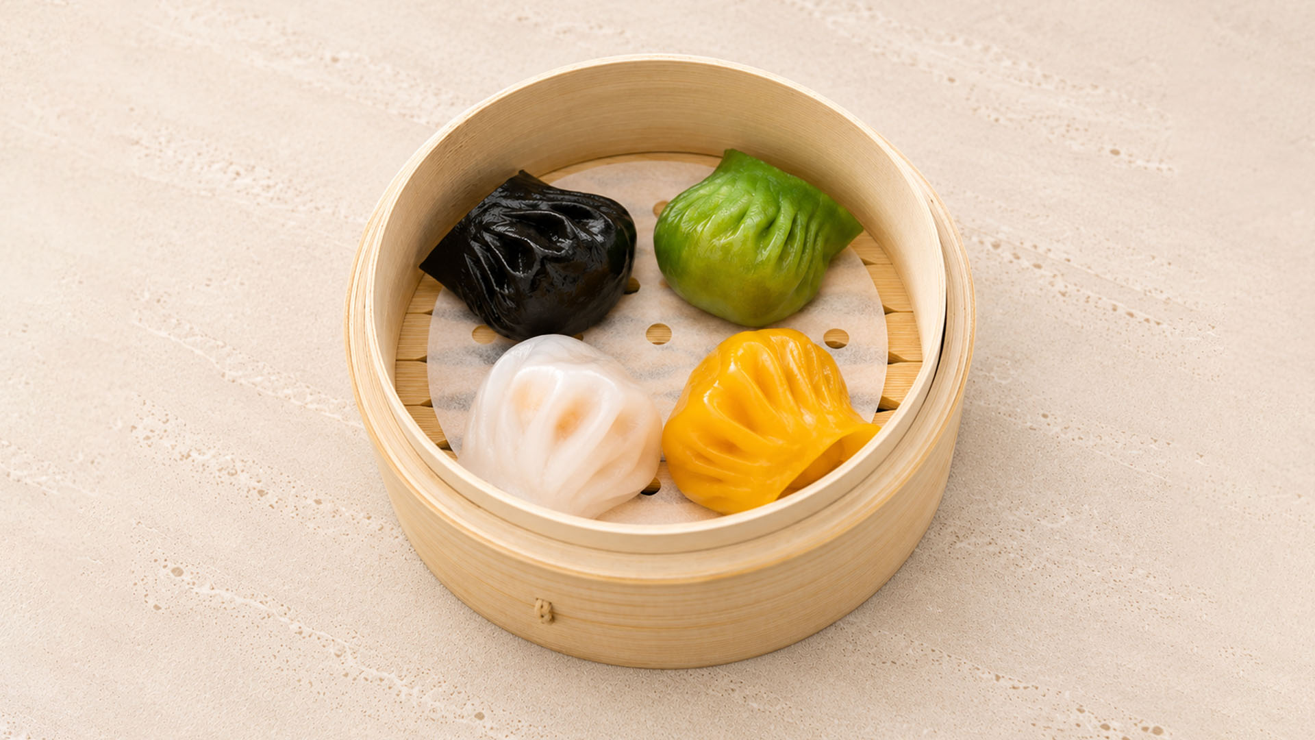 Four steamed dumplings in black, green, white, and yellow in a bamboo steamer