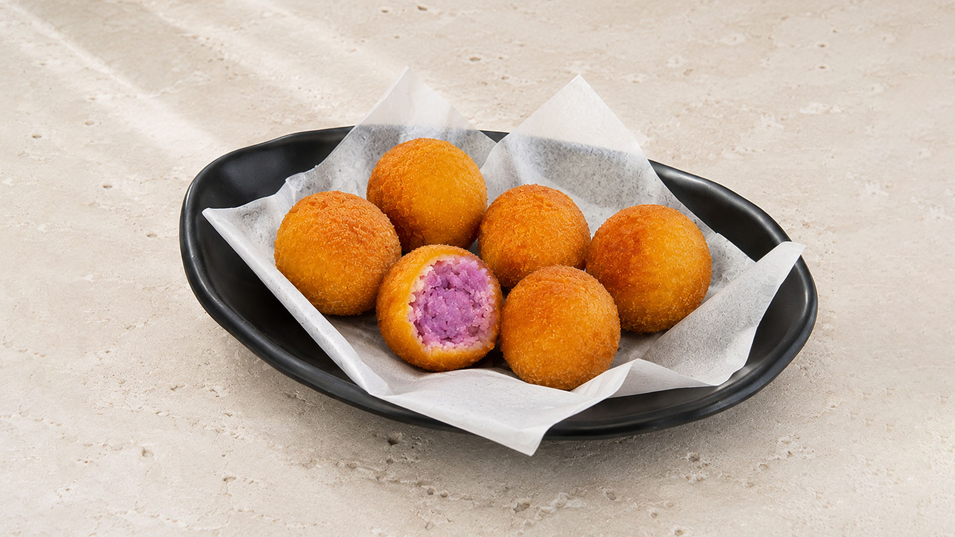 Golden fried purple sweet potato balls, one broken open, on a black plate