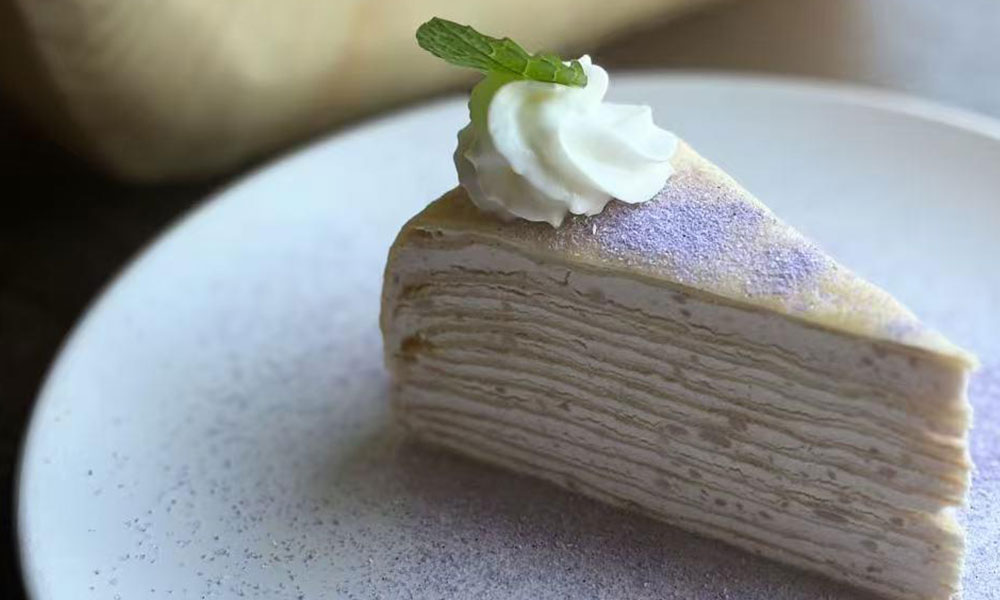 Slice of purple crepe mille cake topped with whipped cream and fresh mint leaf on speckled white plate at Dumplings & Tea, a Asian Restaurant in Chesterfield