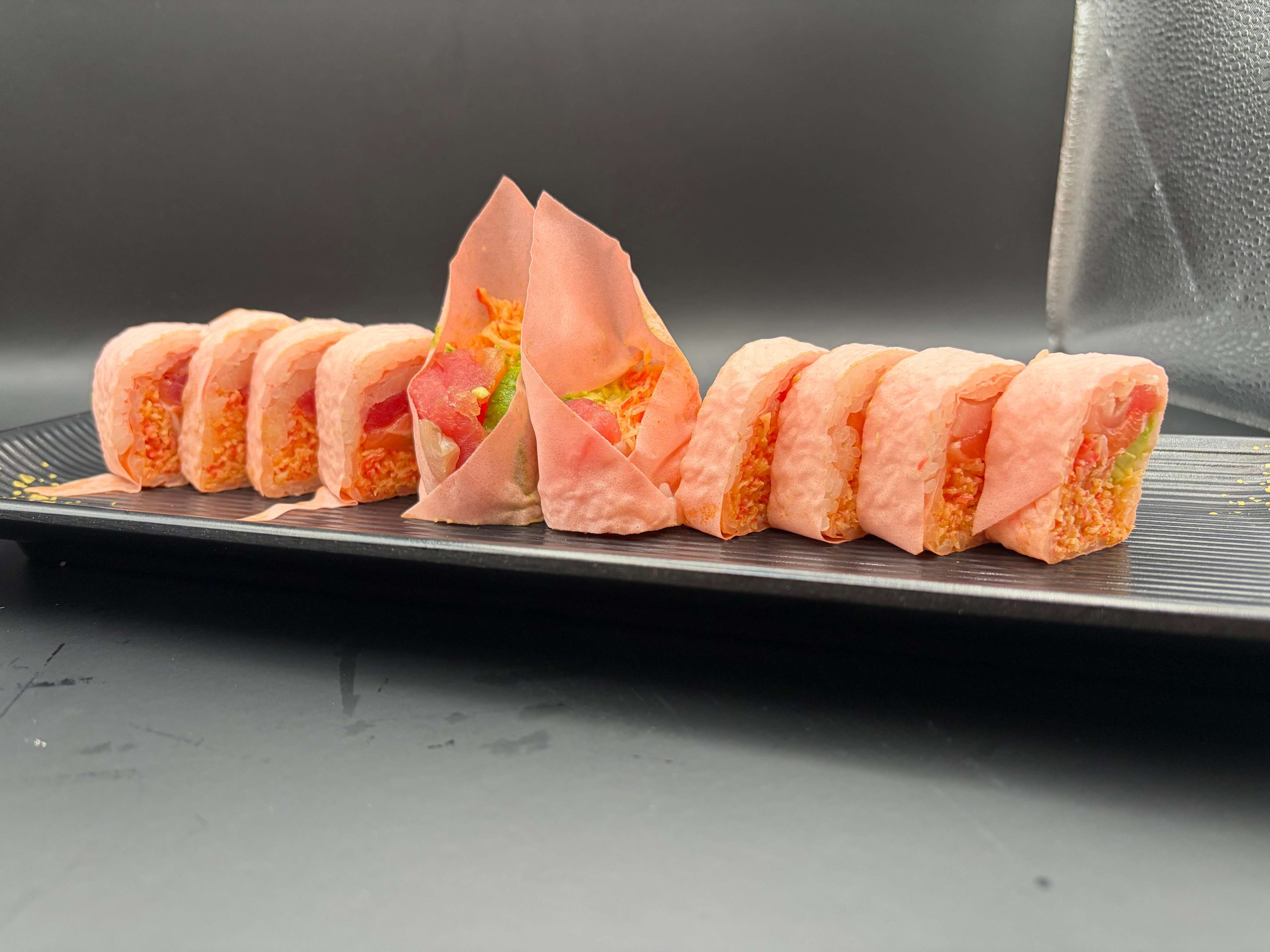 Pink-wrapped sushi roll filled with seafood, avocado, and rice, arranged on a black plate
