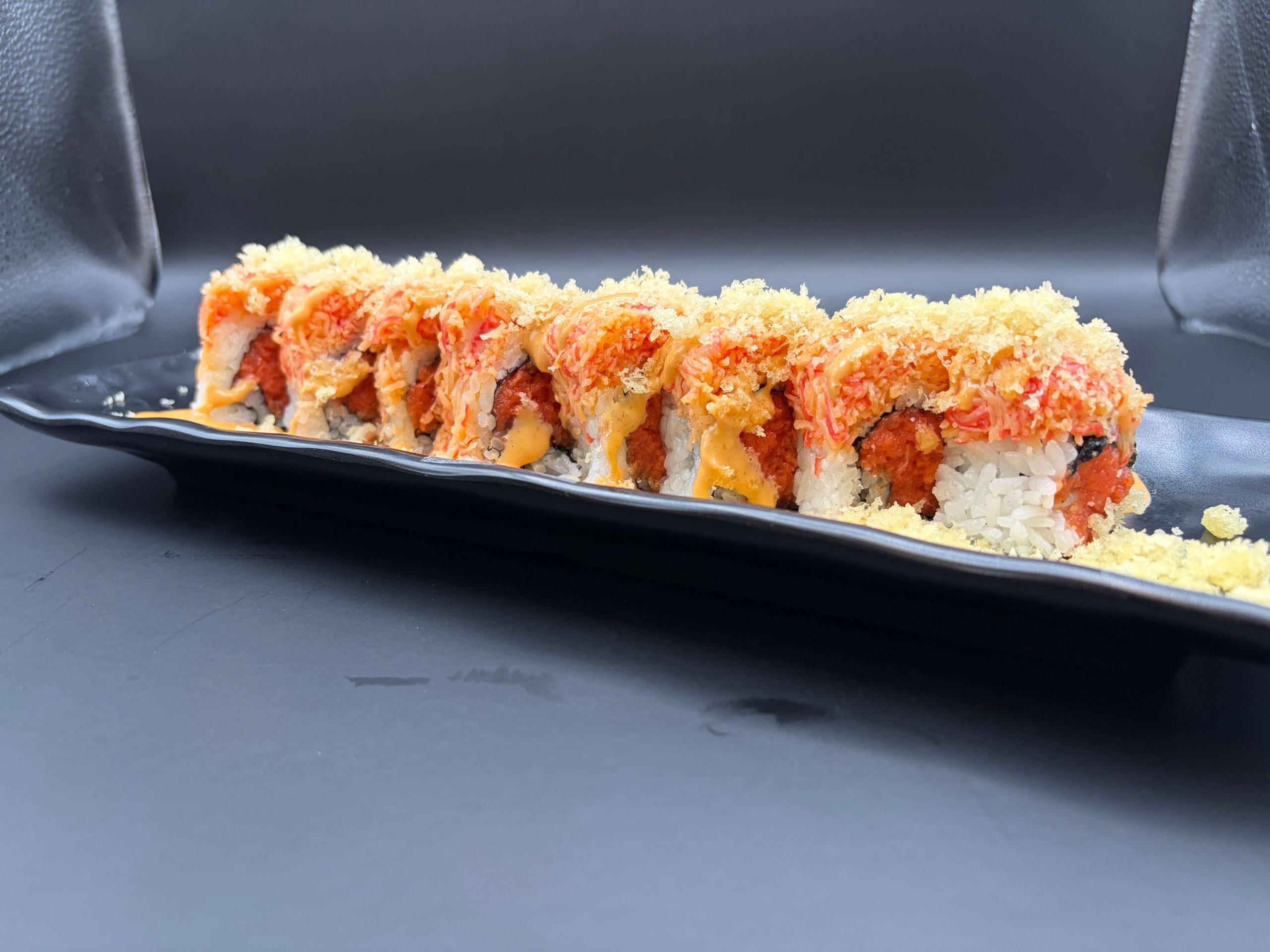 Spicy sushi roll topped with crab mix, crunchy flakes, and creamy sauce on a black plate