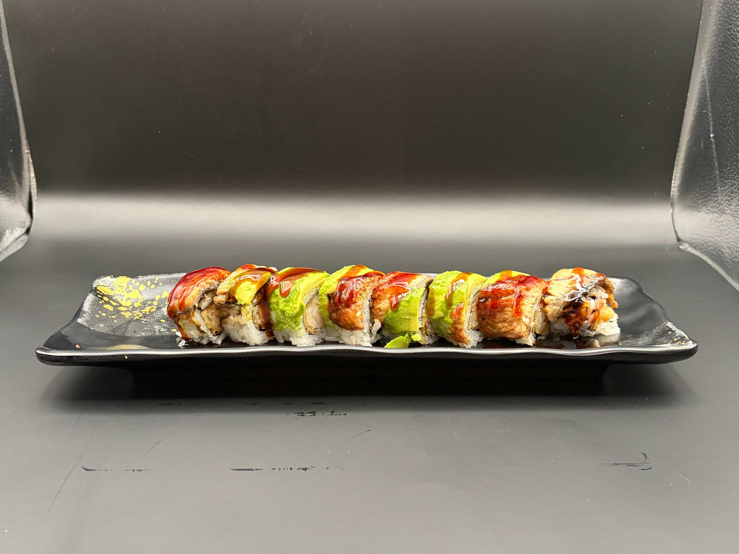Sushi roll topped with avocado, grilled eel, and sweet sauce on a black rectangular plate