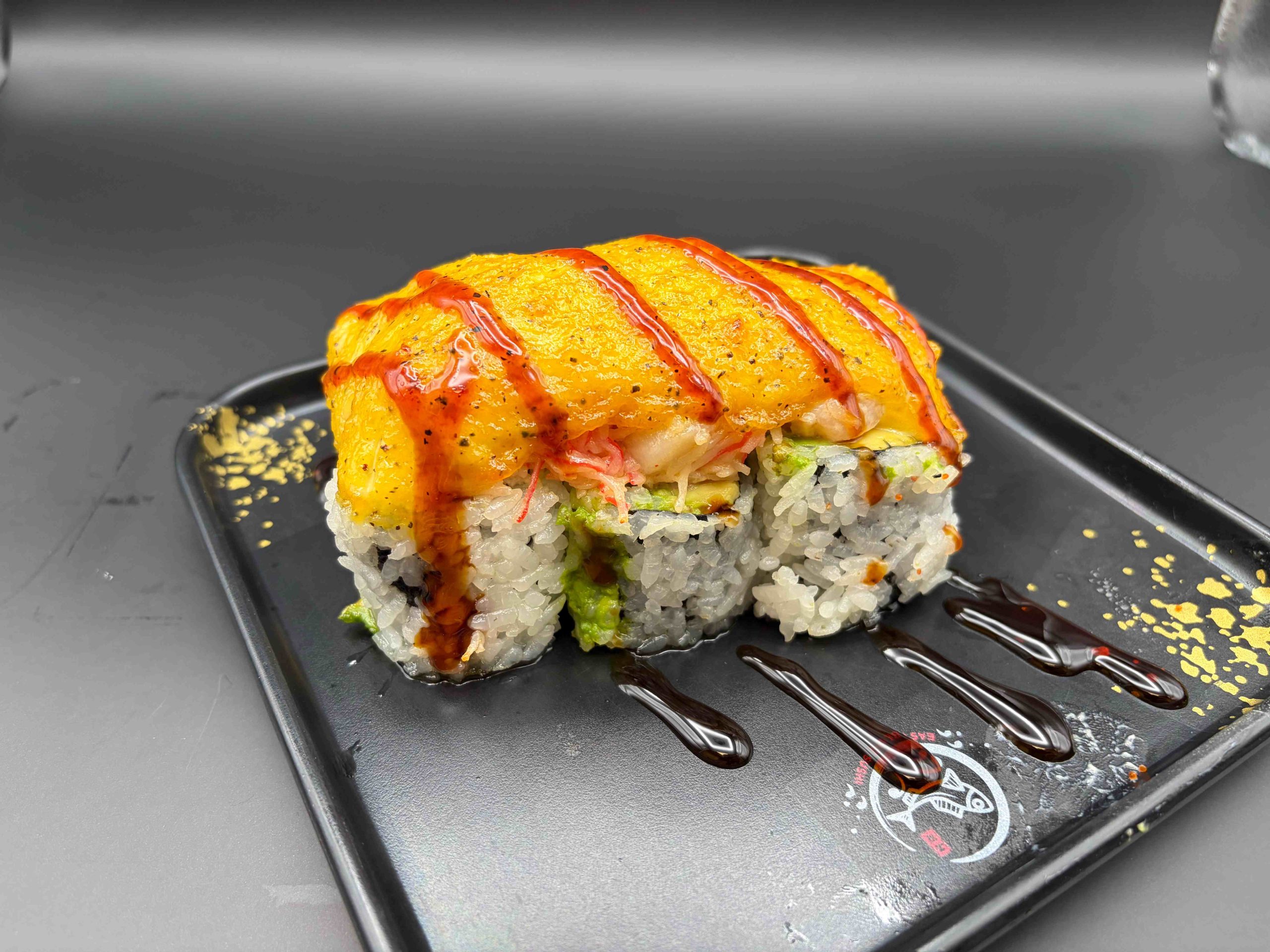 Sushi roll topped with creamy baked seafood, spicy sauce, and sweet glaze on a black plate