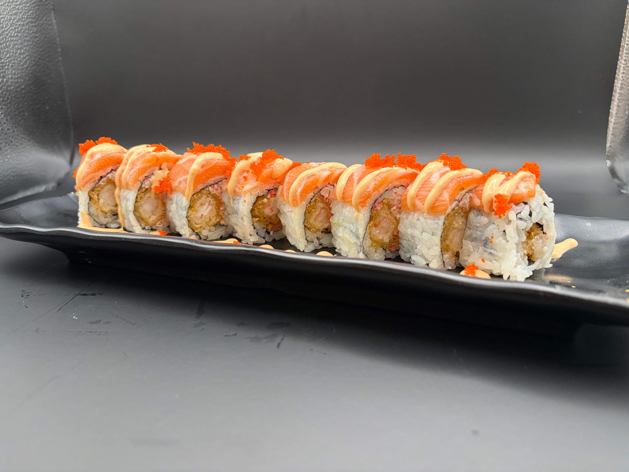 Sushi roll topped with salmon, avocado, shrimp, and sweet sauce on a black plate