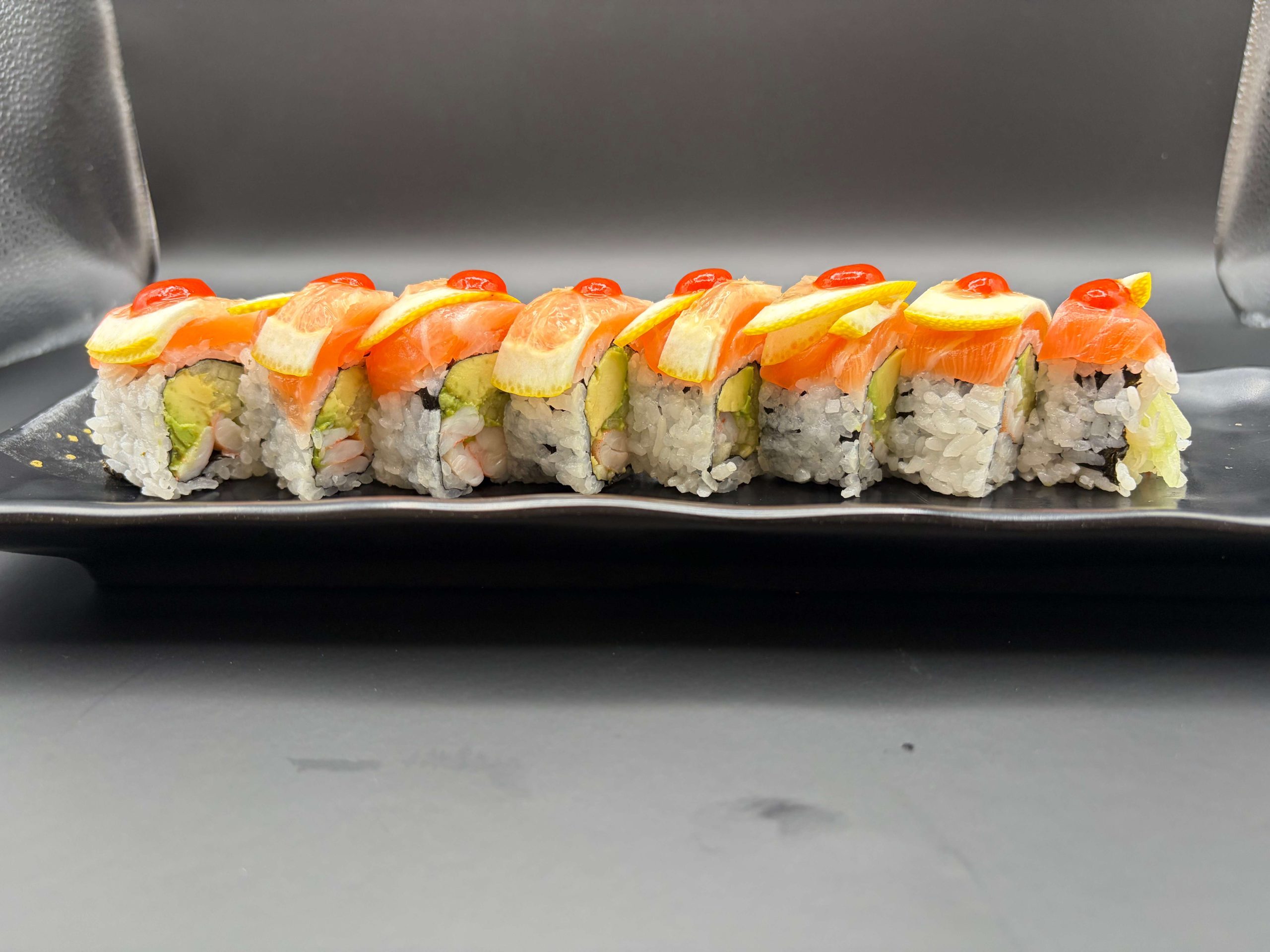 Sushi roll topped with salmon, lemon slices, avocado, and bright red sauce on a black plate