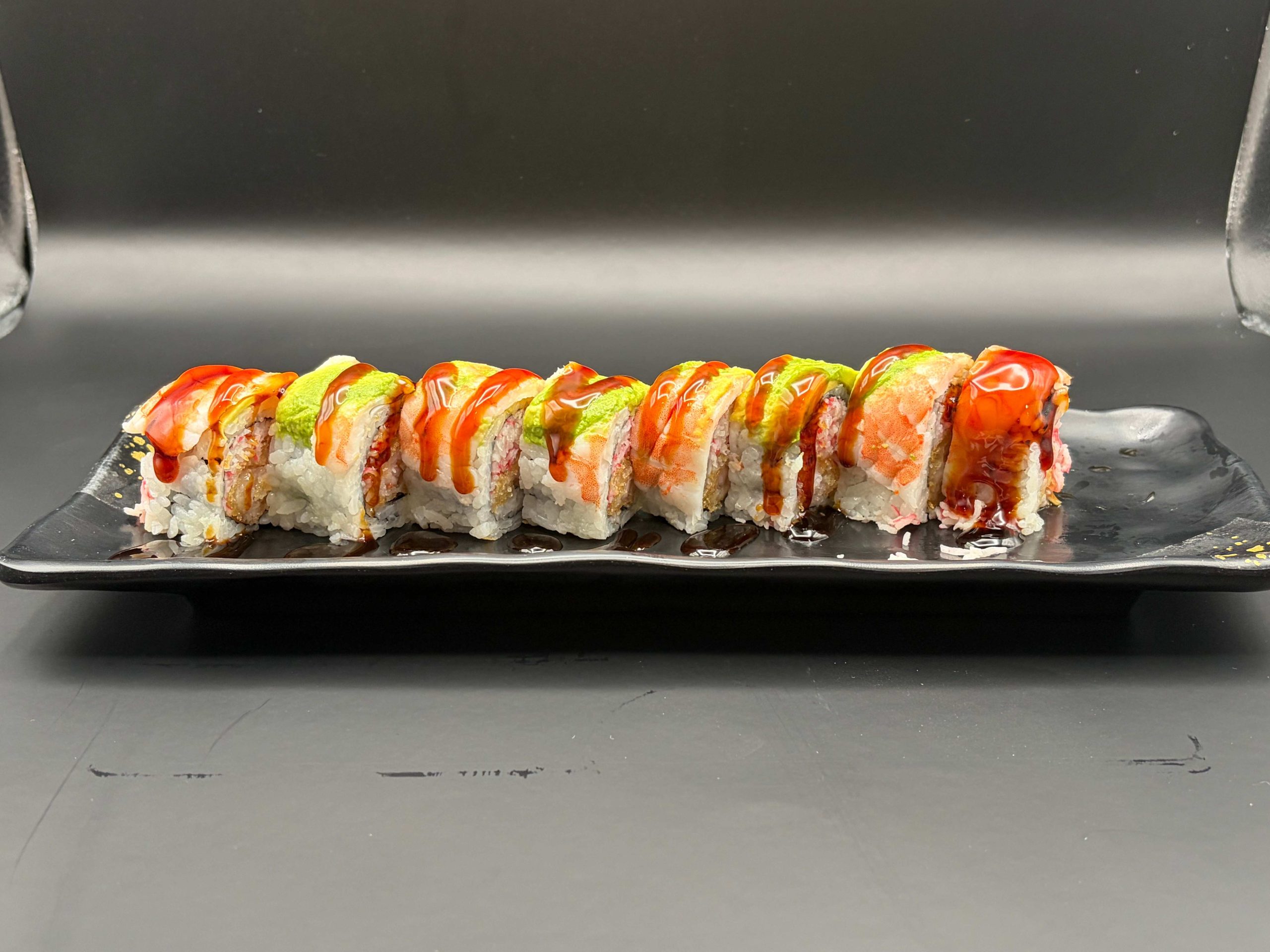 Sushi roll topped with shrimp, avocado, spicy sauce, and sweet glaze on a black plate