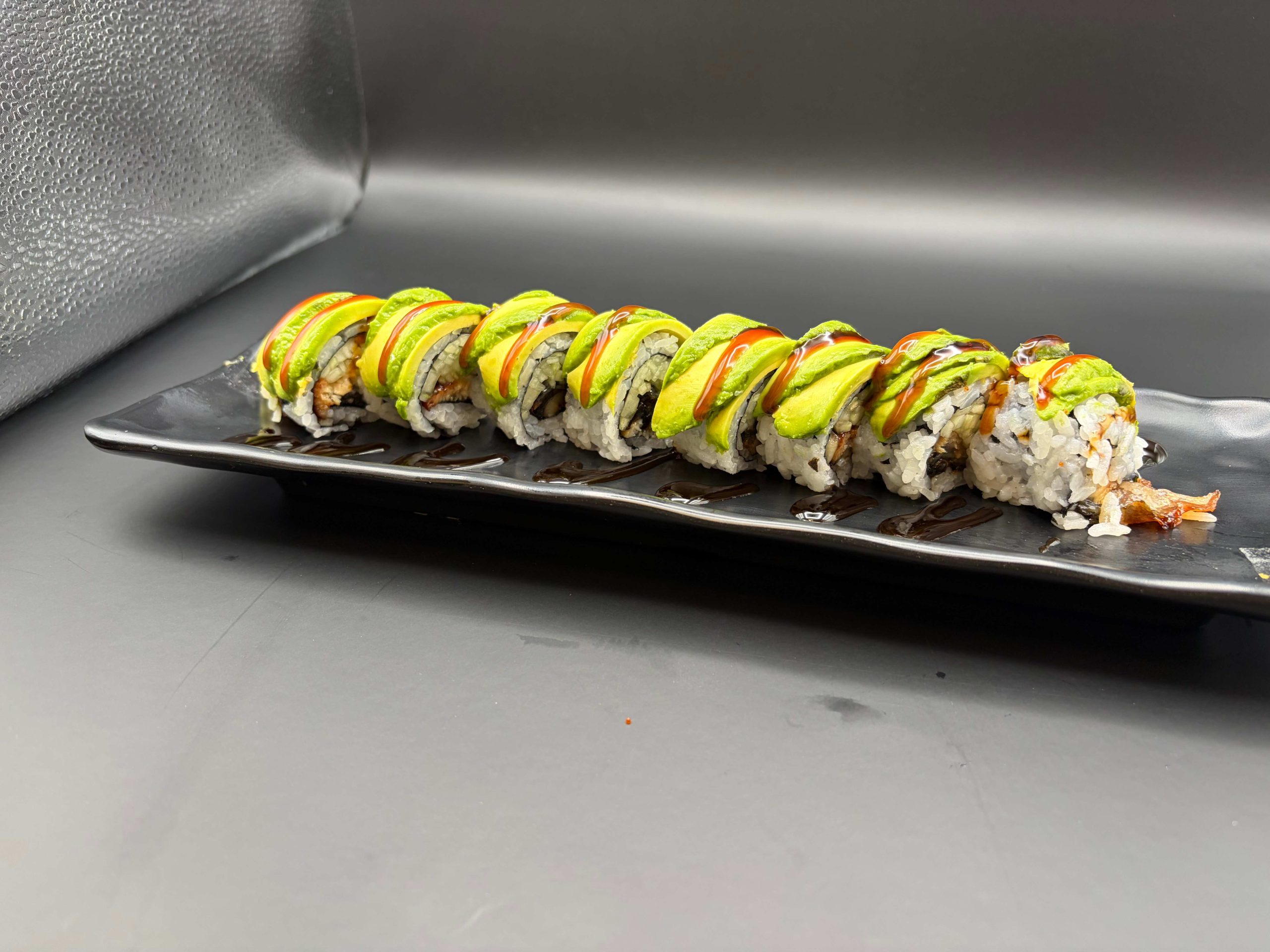 Sushi roll topped with sliced avocado and sweet sauce, served on a black rectangular plate