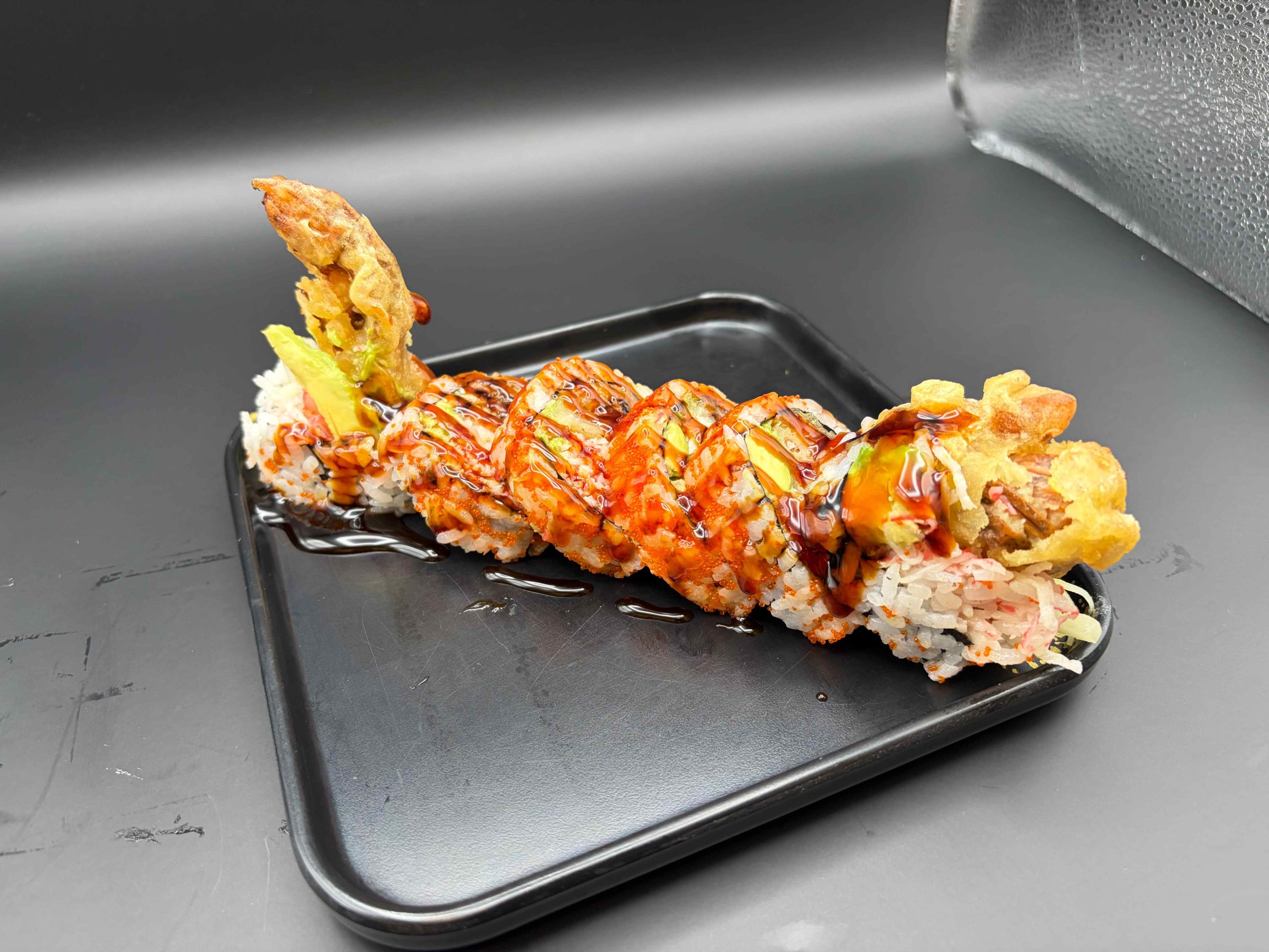 Sushi roll with crispy soft-shell crab, avocado, seafood, and sweet sauce on a black plate