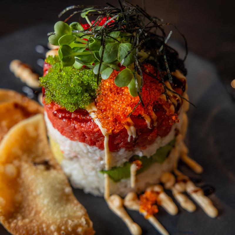 A sushi tower served on a black plate, layered with avocado in the center—viewed from above.