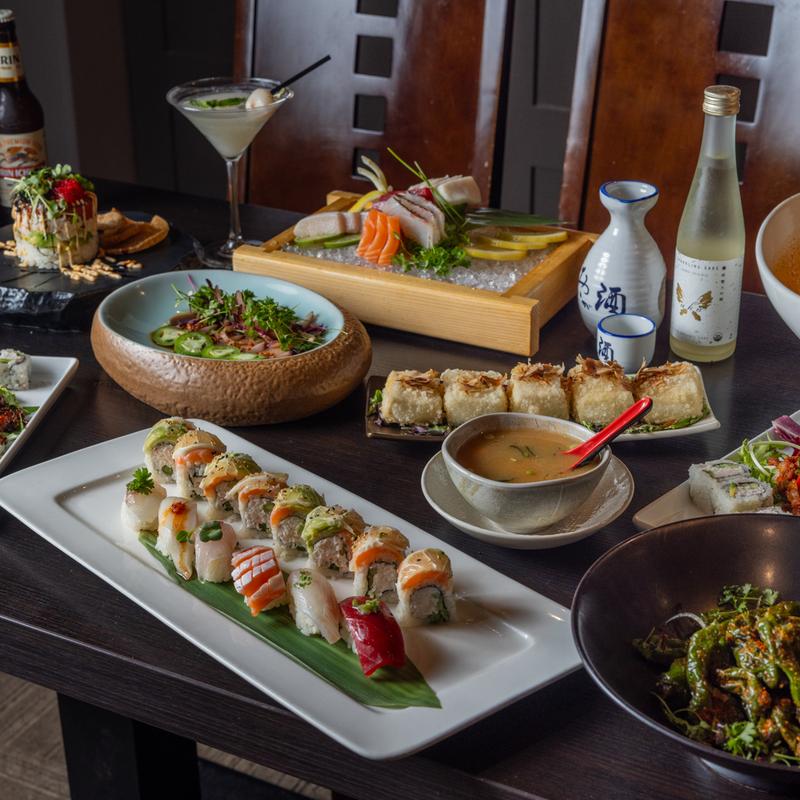 The dark-themed image features a variety of Japanese dishes, along with a bottle of sake.