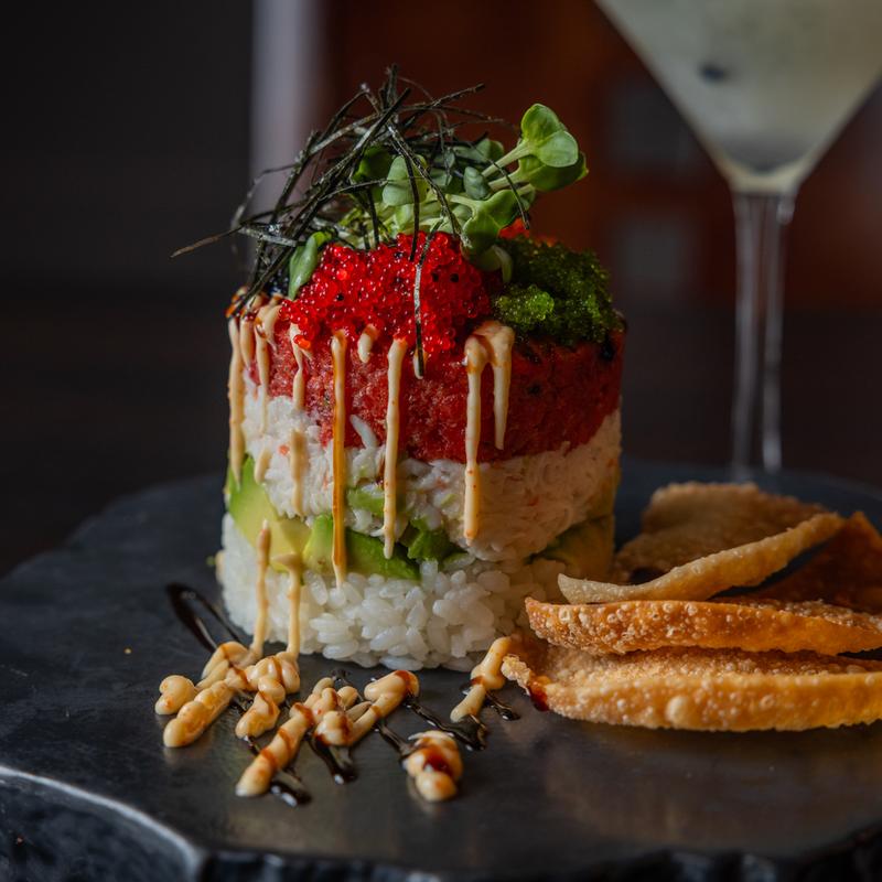 A sushi tower served on a black plate, layered with avocado in the middle.