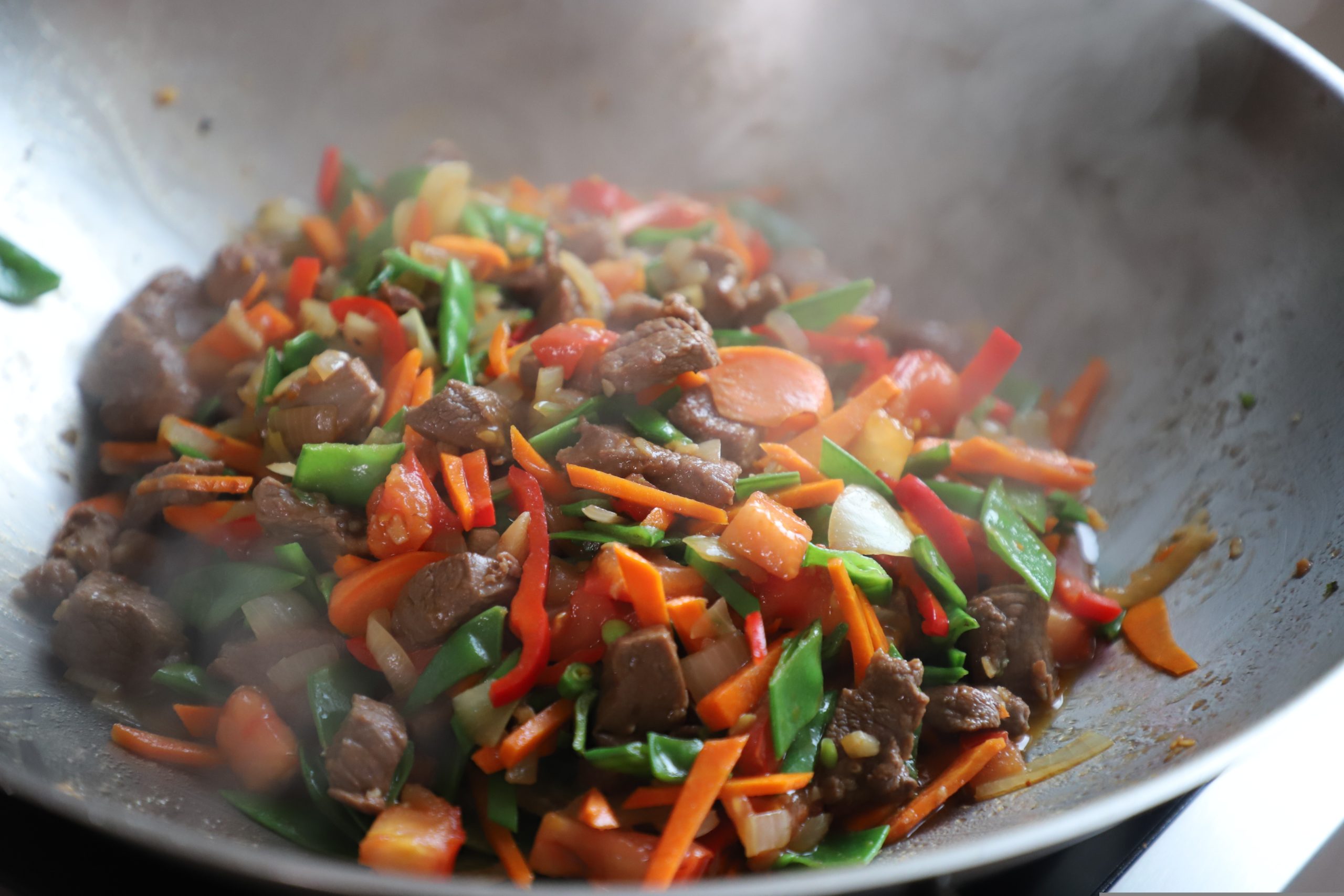 Beef and colorful vegetables being stir-fried in a metal wok with visible steam