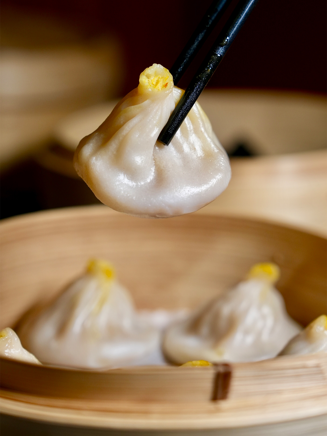 Floral-Chinese-Restaurant-New-York Extreme close-up of a soup dumpling (xiao long bao) held by black chopsticks above a bamboo steamer.