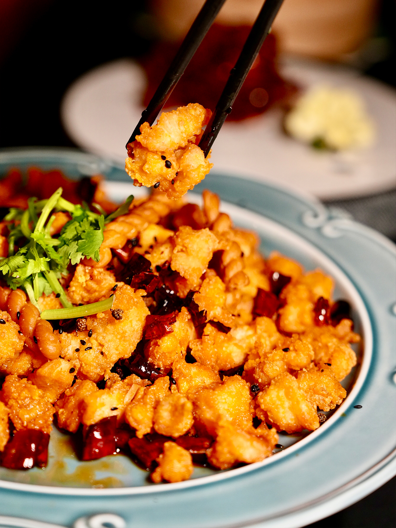 Floral-Chinese-Restaurant-New-York Close-up of Kung Pao chicken or similar spicy, deep-fried chicken dish with chili peppers and peanuts. Chopsticks hold up a piece.