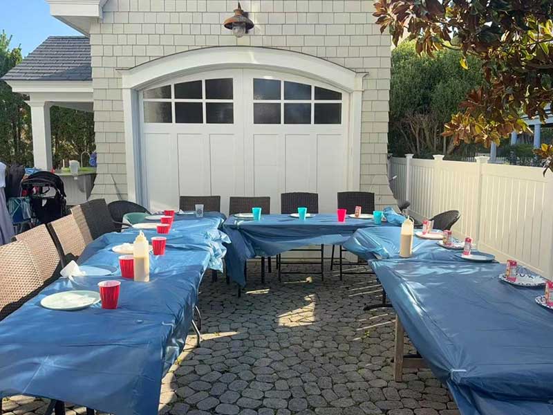 Outdoor patio party dining area, long tables covered with blue plastic tablecloths, red and turquoise disposable cups, plates and squeeze bottles, stone paving, white garage door and residential house background.