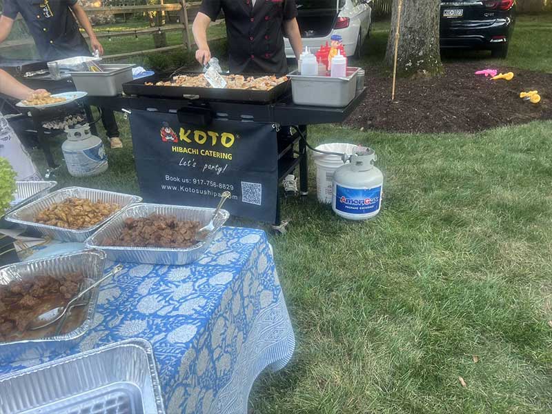 Koto hibachi catering outdoor event, chefs cook food on portable grill, aluminum foil trays with prepared meats on table, propane gas tanks, branded booth banner, grass lawn with parked cars in background.