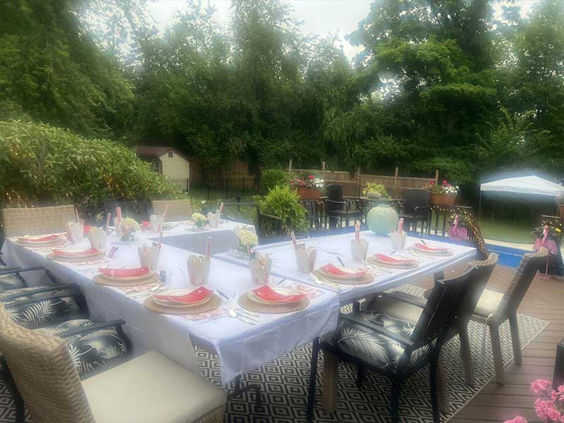 Elegant backyard event dining tables laid with white tablecloths, red napkins, flower centerpieces, dark wicker chairs, wooden deck, dense green trees, potted blooms and distant pool area.