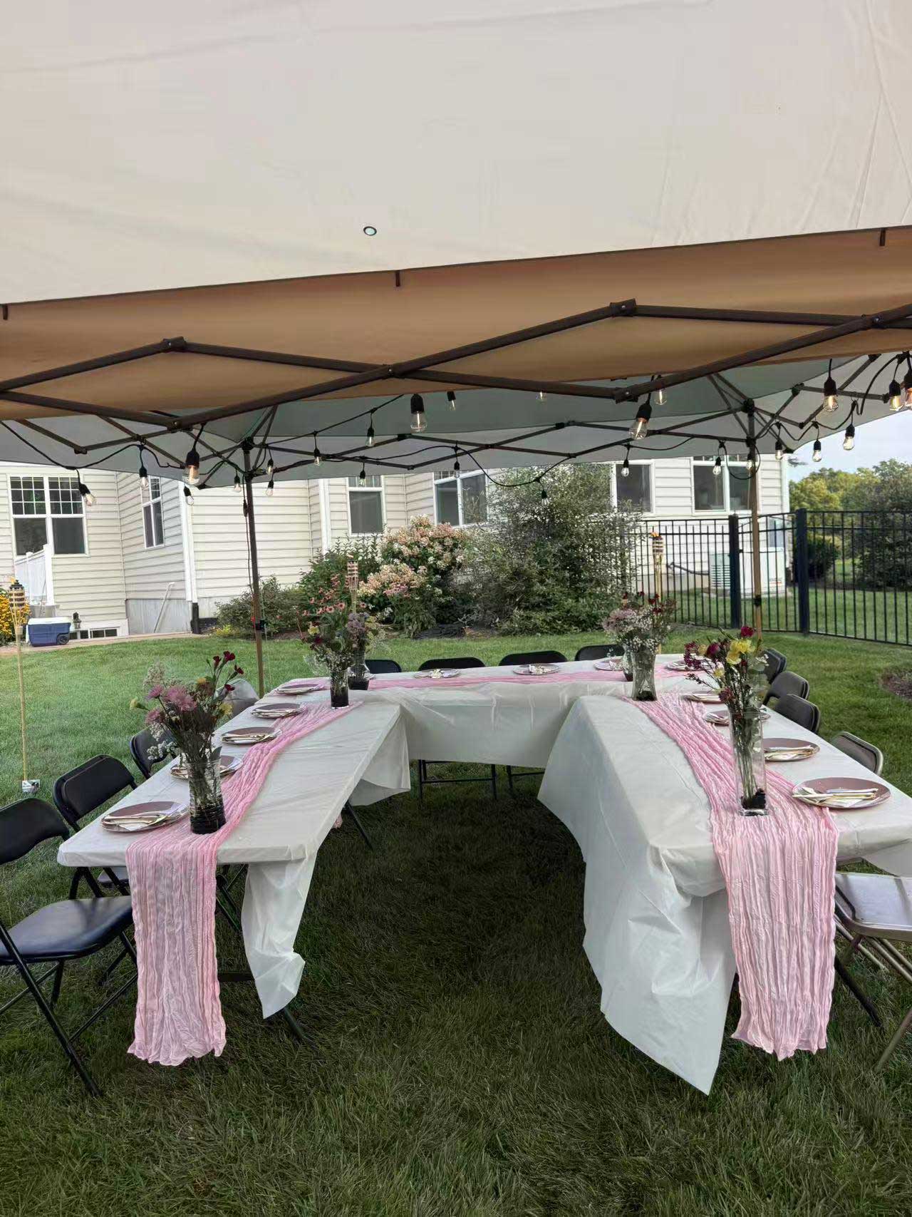 Backyard party dining setup under cream pop‑up canopy with string lights. White‑covered tables accented by pink fabric runners, wildflower vase centerpieces, place settings, black folding chairs, green lawn and residential house backdrop.
